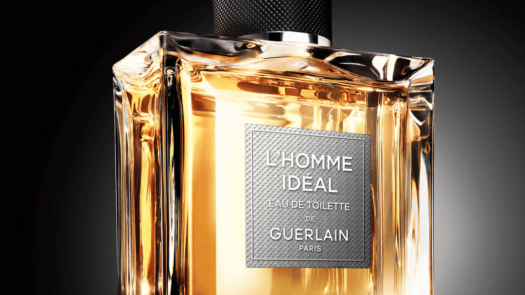 Närbild på Guerlains parfymflaska "L'Homme Ideal Eau de Toilette" med en rektangulär design i tjockt glas och en gyllene vätska. Grå etikett med vit text, mörk bakgrund som framhäver flaskans eleganta och lyxiga utseende.