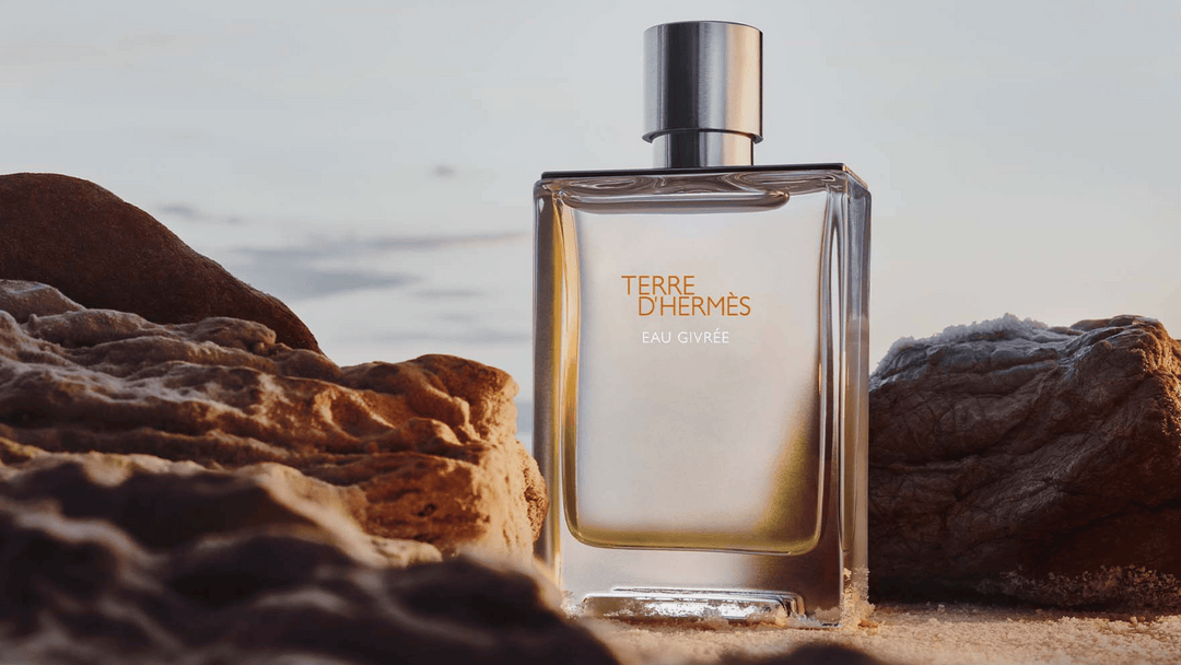 Bilden visar en parfymflaska från Hermès, "Terre D'Hermès Eau Givrée". Flaskan är elegant och minimalistisk, placerad mellan två stenar på en sandstrand, vilket ger en naturlig och jordnära känsla. 