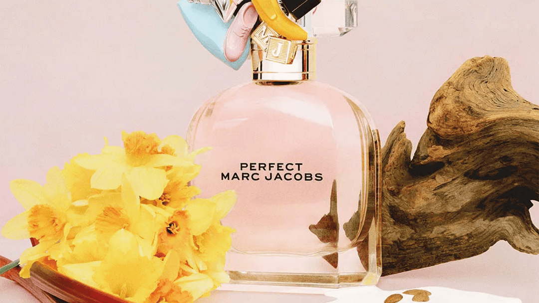 På bilden ser vi en parfymflaska från Marc Jacobs, märkt som "Perfect". Flaskan är omgiven av en lekfull och konstnärlig arrangemang, inklusive livfulla gula blommor och en intrikat träbit, vilket ger en naturlig och organisk känsla till presentationen.