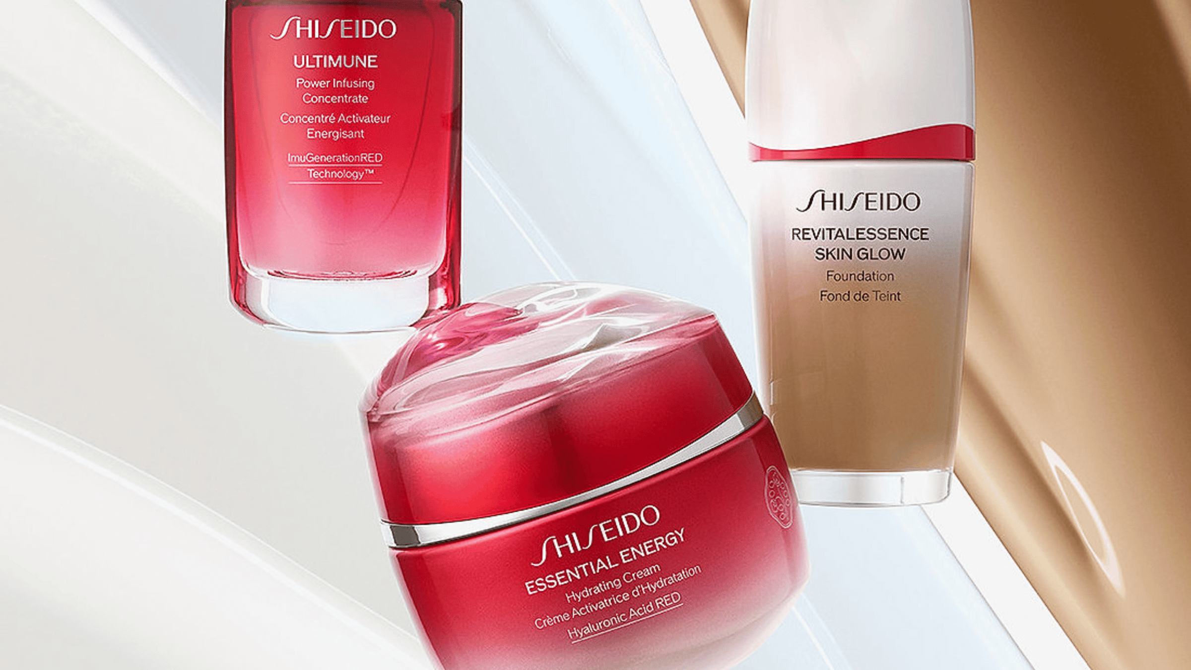 En samling av Shiseido-produkter, inklusive Ultimune Power Infusing Concentrate, Essential Energy Hydrating Cream och Revitalessence Skin Glow Foundation. Produkterna har eleganta förpackningar i rött och vitt, placerade mot en mjukt belyst bakgrund.