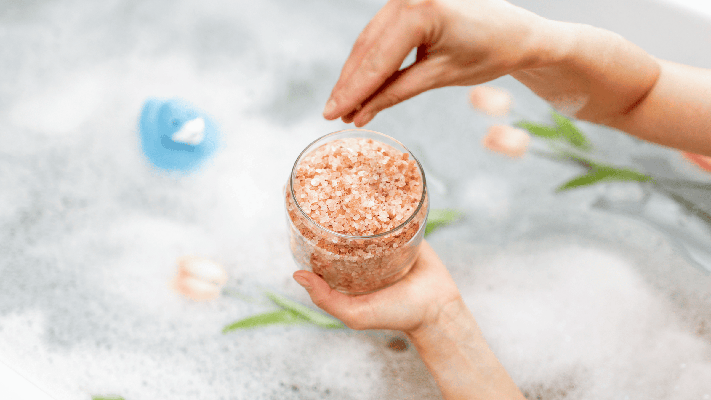 En hand håller ett genomskinligt glas fyllt med rosa badsalt, redo att hällas i ett bubbelbad. I bakgrunden syns en suddig leksaksanka och gröna växter, vilket tillför en lugn och avslappnad atmosfär.