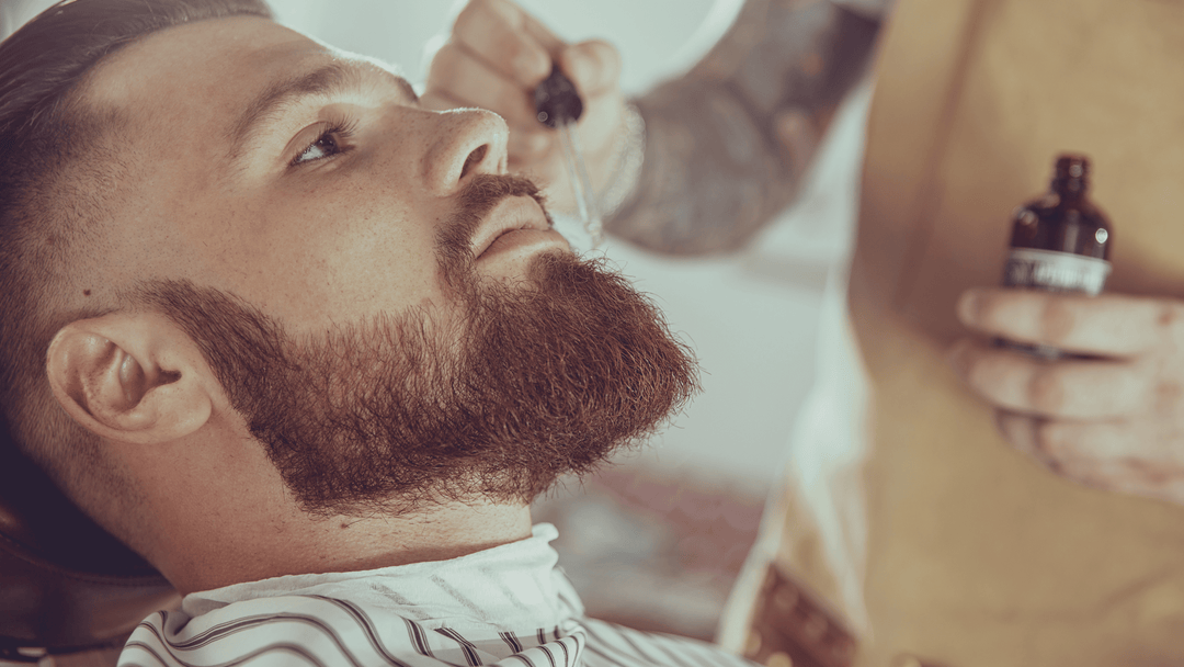 En man ligger tillbakalutad i en barberarstol medan en barberare applicerar skäggolja på hans välvårdade skägg. Barberaren är klädd i ett beige förkläde och arbetar omsorgsfullt, vilket skapar en professionell och avslappnad atmosfär.